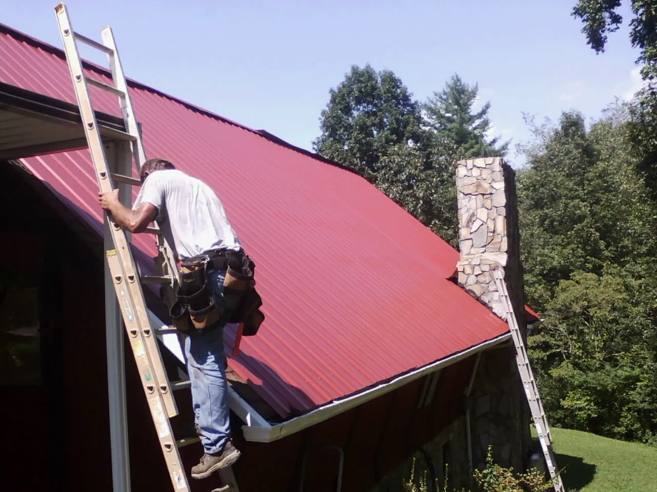 Roofer on a ladder beside a red metal roof and stone chimney — Soffit & Fascia Repair by Roofing Brothers in Kingsport